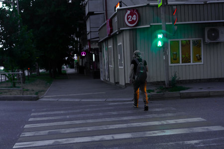 A person with a backpack walks across a crosswalk while a green signal glows. Nearby buildings and trees create a calm atmosphere during early evening.の写真素材