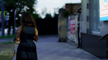 A woman walks down a dimly lit street, surrounded by urban architecture. The evening sky casts soft shadows, creating a tranquil atmosphere as she moves along the pavement.の写真素材