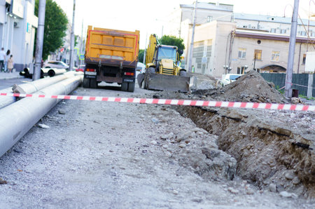Excavation activities are in progress on a city road with construction vehicles parked nearby. The area is marked with safety tape, indicating work in progress during the late afternoon.の写真素材