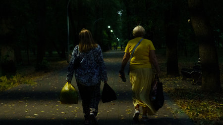 Two women stroll down a dimly lit park pathway, holding bags as fallen leaves surround them. The atmosphere is calm, typical of a serene evening walk.の写真素材