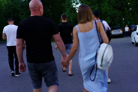A group of friends strolls down a tree-lined street during the evening. They chat and hold hands,...