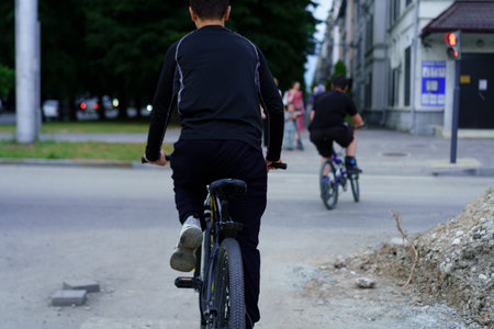 Two young cyclists ride their bikes as dusk falls on a city street. The surroundings include urban buildings and trees, creating a vibrant evening atmosphere.の写真素材