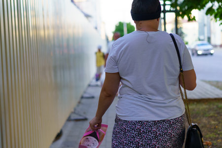 A woman strolls down a busy urban sidewalk, holding colorful posters. Construction barriers line the path as she enjoys the sunny weather and vibrant city life.の写真素材