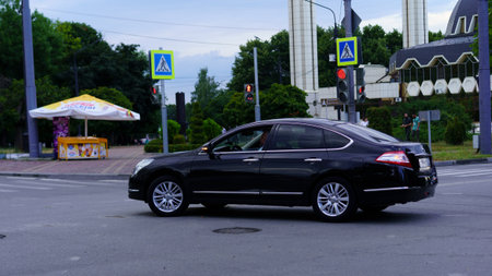 A black sedan is making a turn at an intersection, surrounded by traffic lights and pedestrian crossing signs. It is a cloudy afternoon in an urban area with green trees.の写真素材