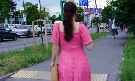 A woman dressed in a pink sundress strolls along a city sidewalk on a summer evening. The scene features nearby cars and pedestrians, creating a lively atmosphere.の写真素材