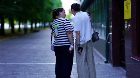 Two friends share a friendly conversation while walking hand in hand on a peaceful street surrounded by lush trees in the afternoon sunlight.の写真素材