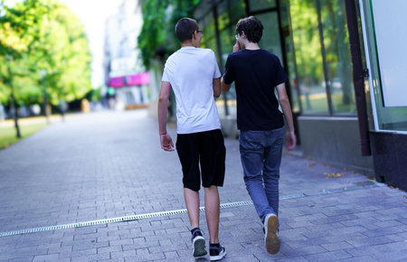 Two young men casually stroll side by side on a vibrant urban street, enjoying the warm summer weather and each other's company amidst green trees.の写真素材