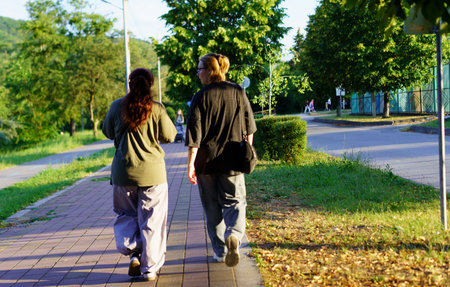 Two individuals stroll down a pathway in a park, surrounded by lush greenery under bright sunlight. The atmosphere is calm and serene as they engage in conversation.の写真素材