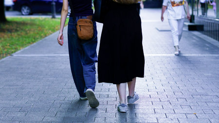 Two friends stroll side by side along a paved path in a city park, enjoying a sunny afternoon, amidst greenery and light chatter from nearby pedestrians.の写真素材
