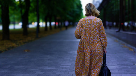 A woman dressed in a flowery yellow dress walks leisurely along a cobblestone path surrounded by trees, with soft evening light illuminating the scene.の写真素材