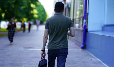 A man walks along a bustling city sidewalk, holding a small bag in one hand. The trees cast shadows...