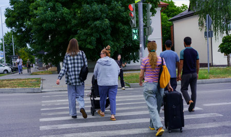 A group of people walks across a crosswalk near a stoplight in the city. The scene captures a mix of individuals, including a family with a stroller and travelers with luggage.の写真素材