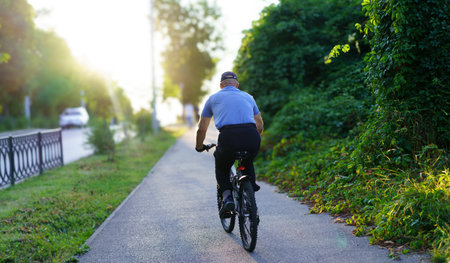 An elderly man rides a bicycle on a serene path surrounded by lush greenery. The warm glow of sunset enhances the peaceful atmosphere of the suburban neighborhood.の写真素材