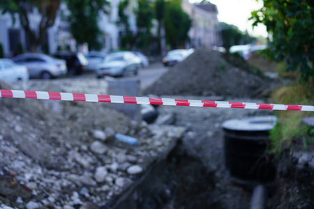 A busy urban street is undergoing construction, featuring a trench filled with gravel and tools. Caution tape visibly restricts access, while vehicles are parked on the road nearby.の写真素材