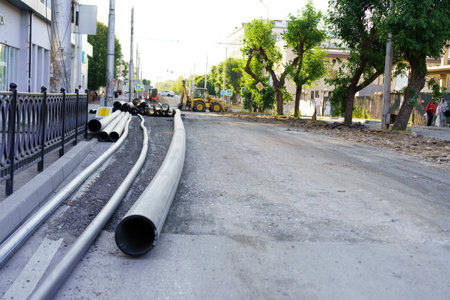 A busy city street is being renovated with large pipes laid out along the side. Heavy machinery works on the road while trees line the street under a clear sky.の写真素材