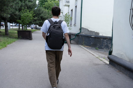 A young individual is seen walking away along a smooth pathway in a city environment, carrying a backpack and surrounded by greenery and buildings under clear skies.の写真素材