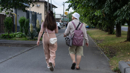 A young woman and an elderly person stroll down a peaceful street, trees lining the path. They have...