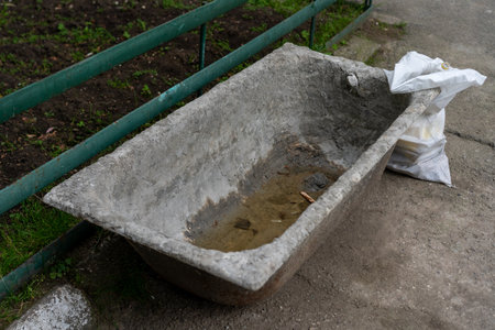 A weathered concrete bathtub is positioned alongside a walkway, filled with stagnant water and debris. A white bag rests nearby, suggesting a forgotten space.の写真素材
