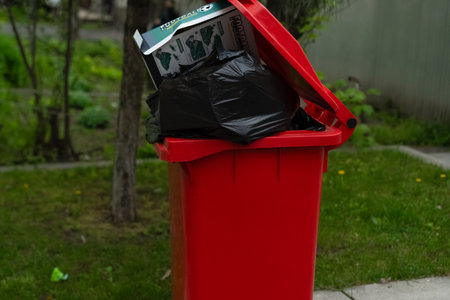 A vibrant red trash bin stands full next to a lush green lawn. The bin is overflowing with black garbage bags and discarded items, indicating a need for collection.の写真素材