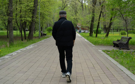 An older man walks alone on a paved path surrounded by lush greenery and trees. The peaceful...
