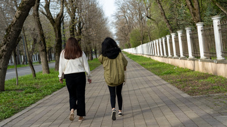 Two women stroll leisurely along a wide, tree-lined walkway in a calm urban neighborhood. Their...