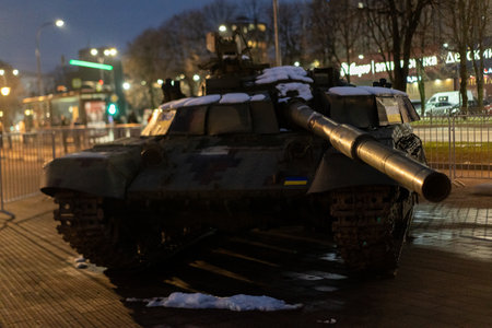 A military tank is positioned in a city square, lit by streetlights, with snow on its surface, symbolizing resilience and history amidst contemporary urban life.の写真素材