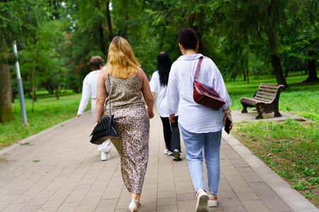 Five friends stroll leisurely along a paved path in a vibrant park, surrounded by greenery and...