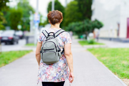 A woman strolls along a city sidewalk, enjoying the sunshine. She wears a floral top and carries a small backpack while passing by greenery and parked cars.の写真素材