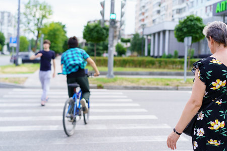 A bicyclist rides through a crosswalk as a man gestures nearby, while a woman walks along the sidewalk. The urban environmental features buildings and greenery.の写真素材