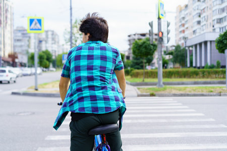 A young individual is seen riding a bicycle while waiting at a crosswalk in an urban environment. The person wears a blue and green checked shirt and looks ahead, surrounded by buildings and traffic.の写真素材