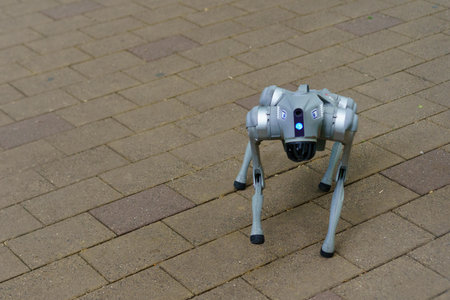 A small, four-legged robot advances along a brick walkway in an urban setting. It is sunny, and the robot features sensors and lights, reflecting advanced technology in robotics.の写真素材
