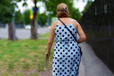 A woman with light brown hair explores her neighborhood, wearing a polka dot dress. She uses a cane for support while strolling past green grass and a black fence on a pleasant day.の写真素材