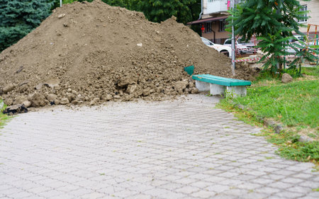 A substantial pile of dirt occupies a park area next to a walkway, while nearby benches provide resting spots for visitors. The scene shows an urban environment during daylight.の写真素材