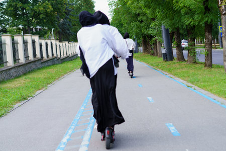 Groups of women, dressed in traditional attire, ride electric scooters on a peaceful pathway surrounded by trees, enjoying a sunny day in the city.の写真素材