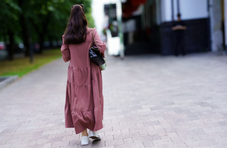 A young woman strolls down a paved path in a park, wearing a long, casual dress and carrying a stylish bag. Green trees line the walkway, creating a serene atmosphere.の写真素材