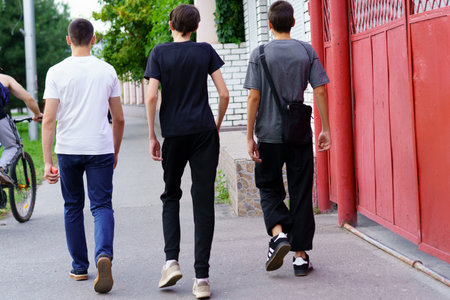 Three teenagers stroll along a sidewalk, enjoying a sunny summer day. They wear casual clothing and...