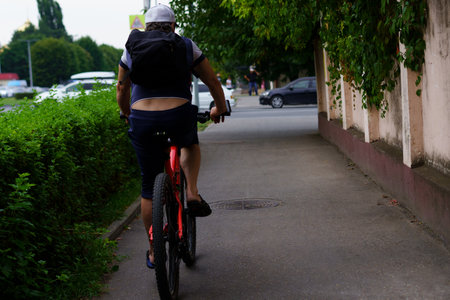 A person on a mountain bike pedals along a city sidewalk bordered by lush plants and parked cars while others walk nearby on a cloudy day.の写真素材