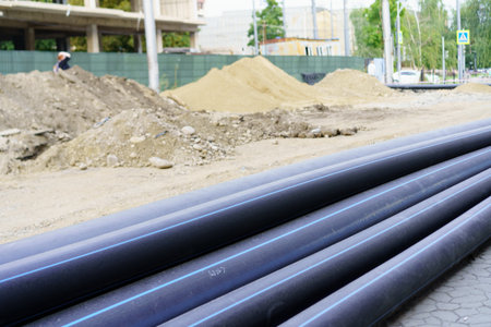 Workers are visible in the background of a construction site. Large black pipes rest on the ground...