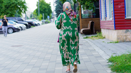 An elderly woman in a vibrant green dress with floral patterns and a matching headscarf strolls...