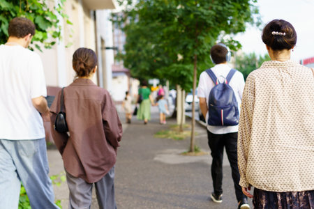 Group of individuals strolling down a sidewalk surrounded by greenery on a clear day. Casual attire indicates a relaxed atmosphere. Lively city scenery can be seen ahead.の写真素材