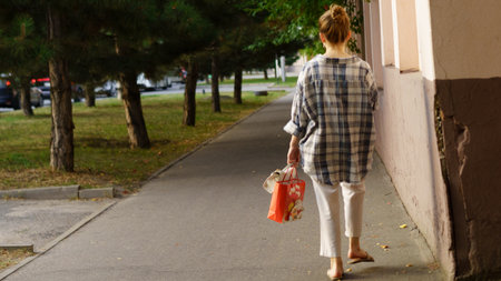 A woman walks barefoot on a sidewalk in a quiet neighborhood, holding a shopping bag. Trees line the...
