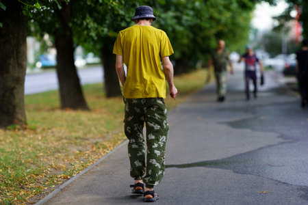 A man wearing a yellow shirt and camo pants walks alone on a sidewalk, while others nearby engage in casual activities. The scene is set in an urban area during daytime.の写真素材