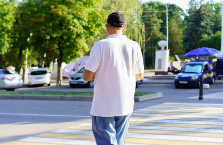 An elderly man walks across a crosswalk during daytime. Surrounding him are parked cars and vendors, creating a lively atmosphere.の写真素材