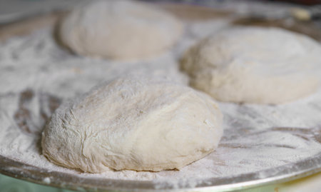 Three pieces of freshly kneaded dough rest on a surface dusted with flour in a cozy kitchen. The...