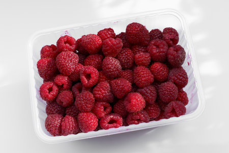 A generous assortment of fresh, plump raspberries fills a transparent container, showing their...