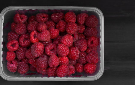 A clear plastic container is filled with vibrant red raspberries, showing their juicy texture and rich color against a dark wooden surface, emphasizing their freshness and appeal.の写真素材