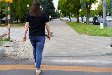 A woman is walking on a paved sidewalk in a city area, surrounded by greenery and parked cars,...