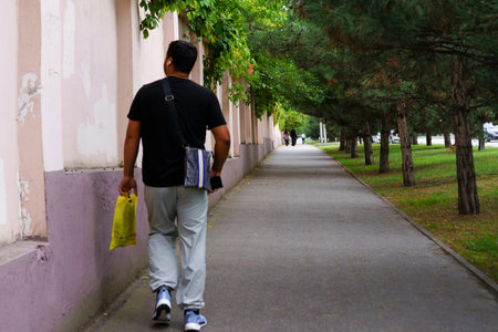 A man strolls down a quiet path surrounded by greenery, holding a yellow shopping bag. The...