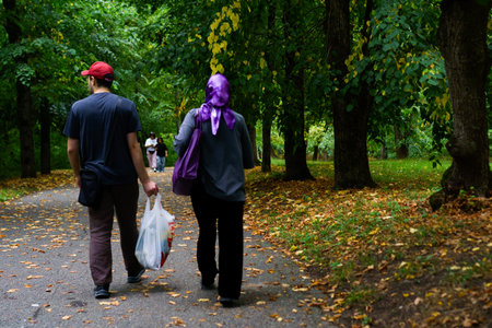 A couple enjoys a tranquil walk in a park filled with autumn leaves. The man carries a bag, and the...
