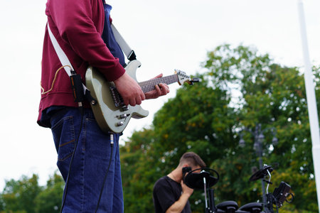 Musicians perform outdoors in a park. One player focuses on his guitar while another adjusts...